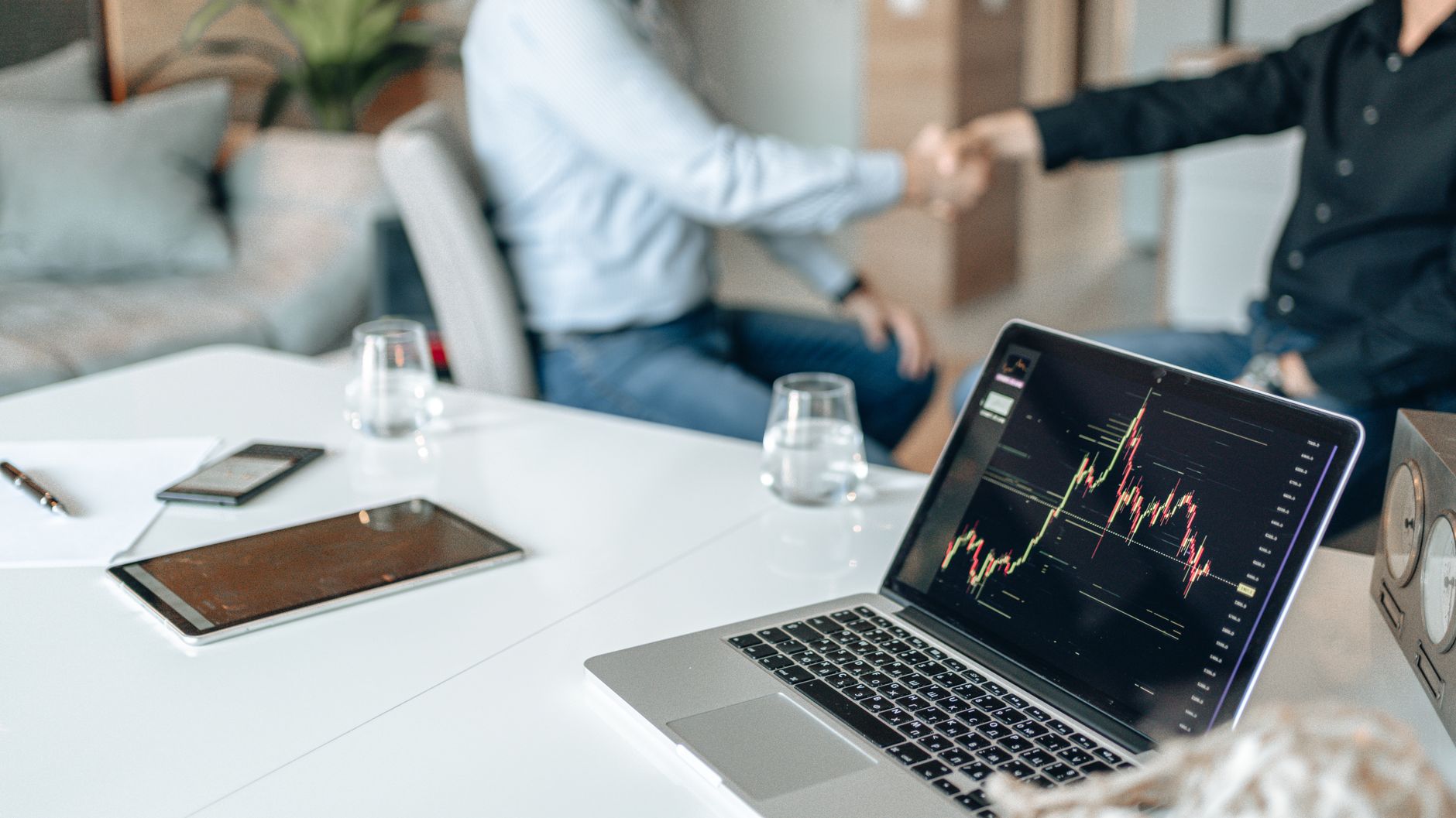 Two professionals shaking hands in an office with data analysis on a laptop screen.