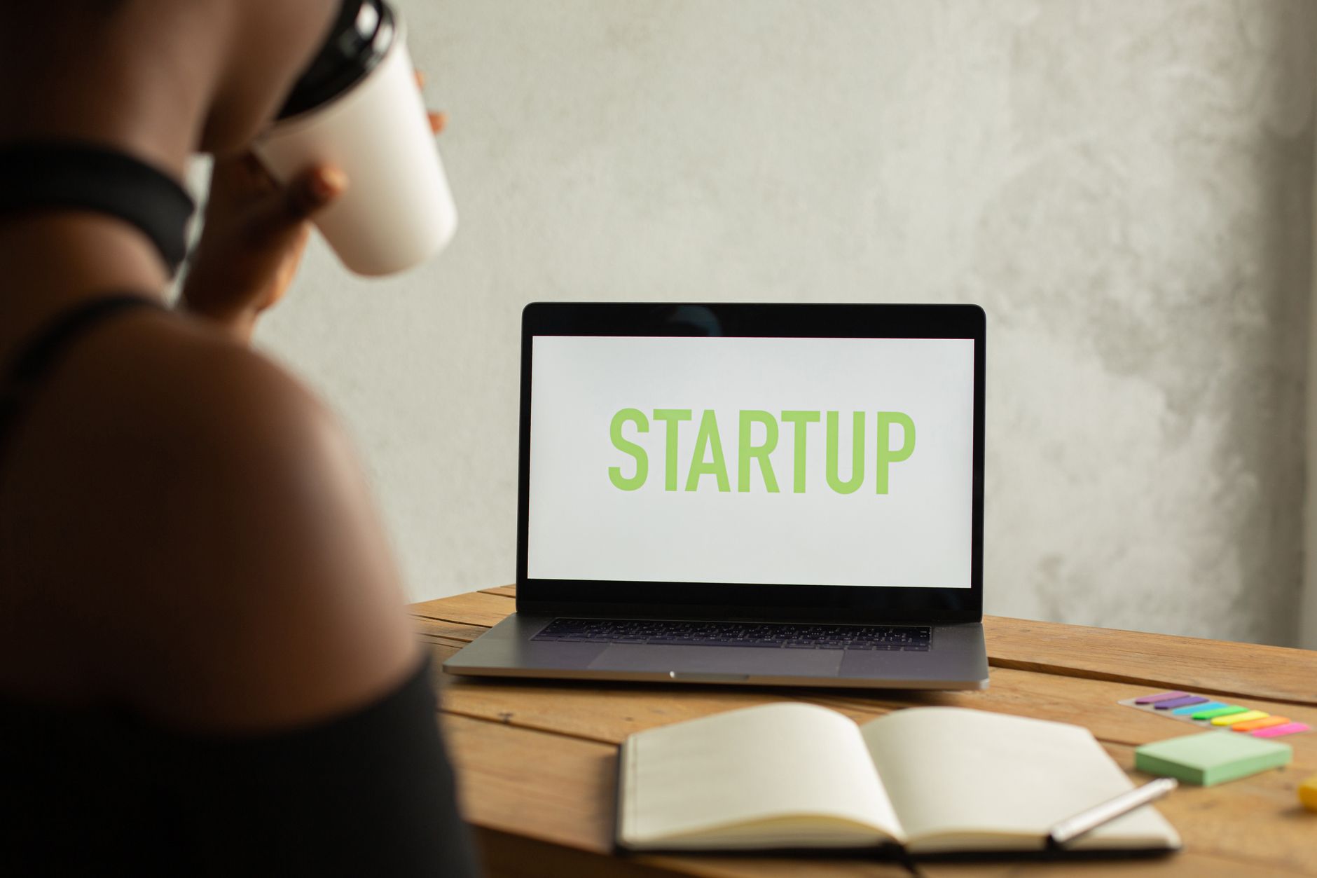 Crop anonymous black female entrepreneur sitting at table with opened netbook with startup on screen and notebook and drinking coffee from takeaway cup while working on project remotely