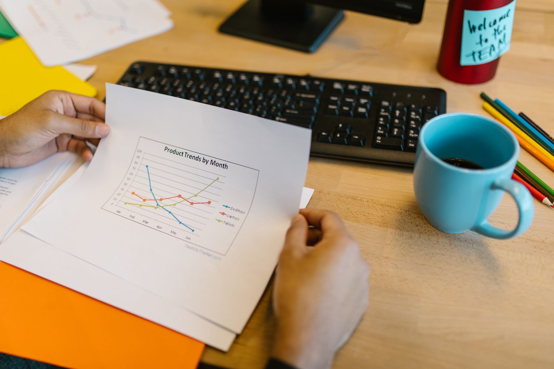 Close-up of hands reviewing a paper graph with a coffee cup and keyboard on a desk.