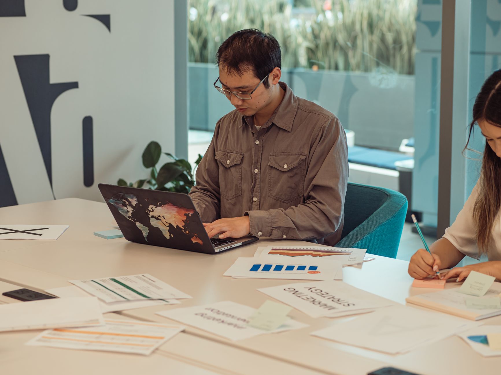 Focused Asian man working on a laptop surrounded by papers in a modern office setting.