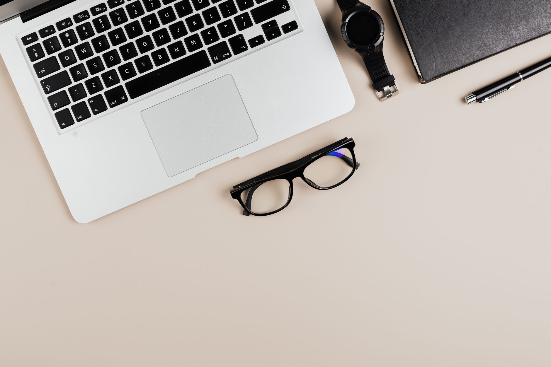 Modern workspace flat lay with laptop, notebook, watch, and glasses on a beige background.