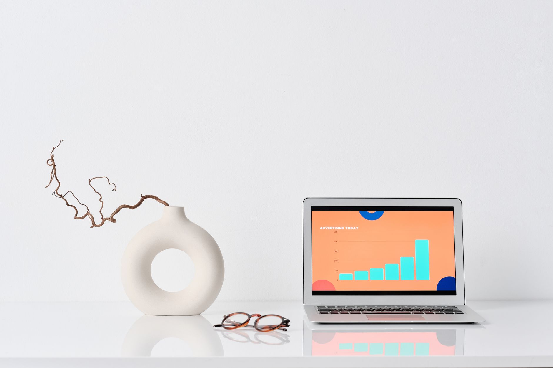 A minimalist workspace with a laptop, ceramic vase, and eyeglasses on a white desk.
