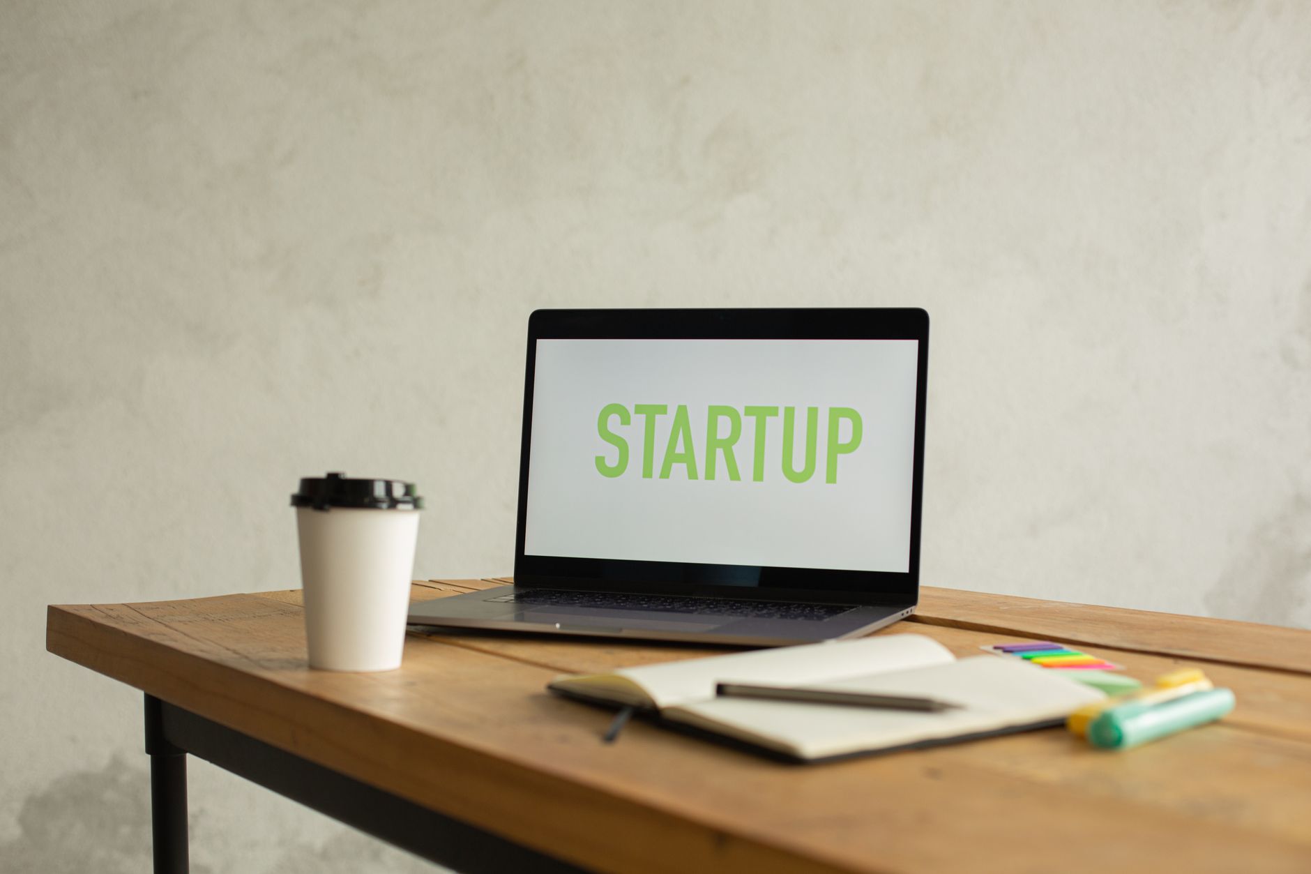 Opened netbook with Startup title on screen and white takeaway cup of coffee placed on wooden table near opened notebook against white wall