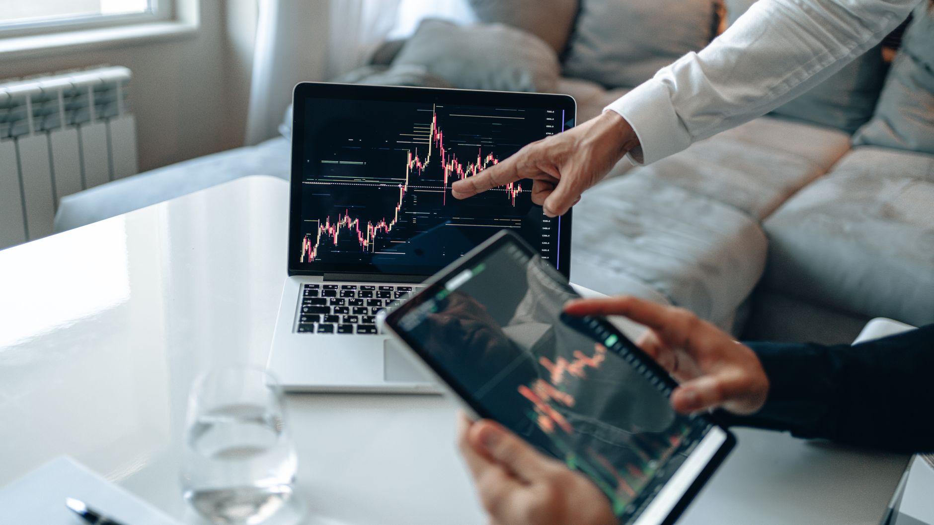 Hands analyzing stock market trends on laptop and tablet, indoors setting.
