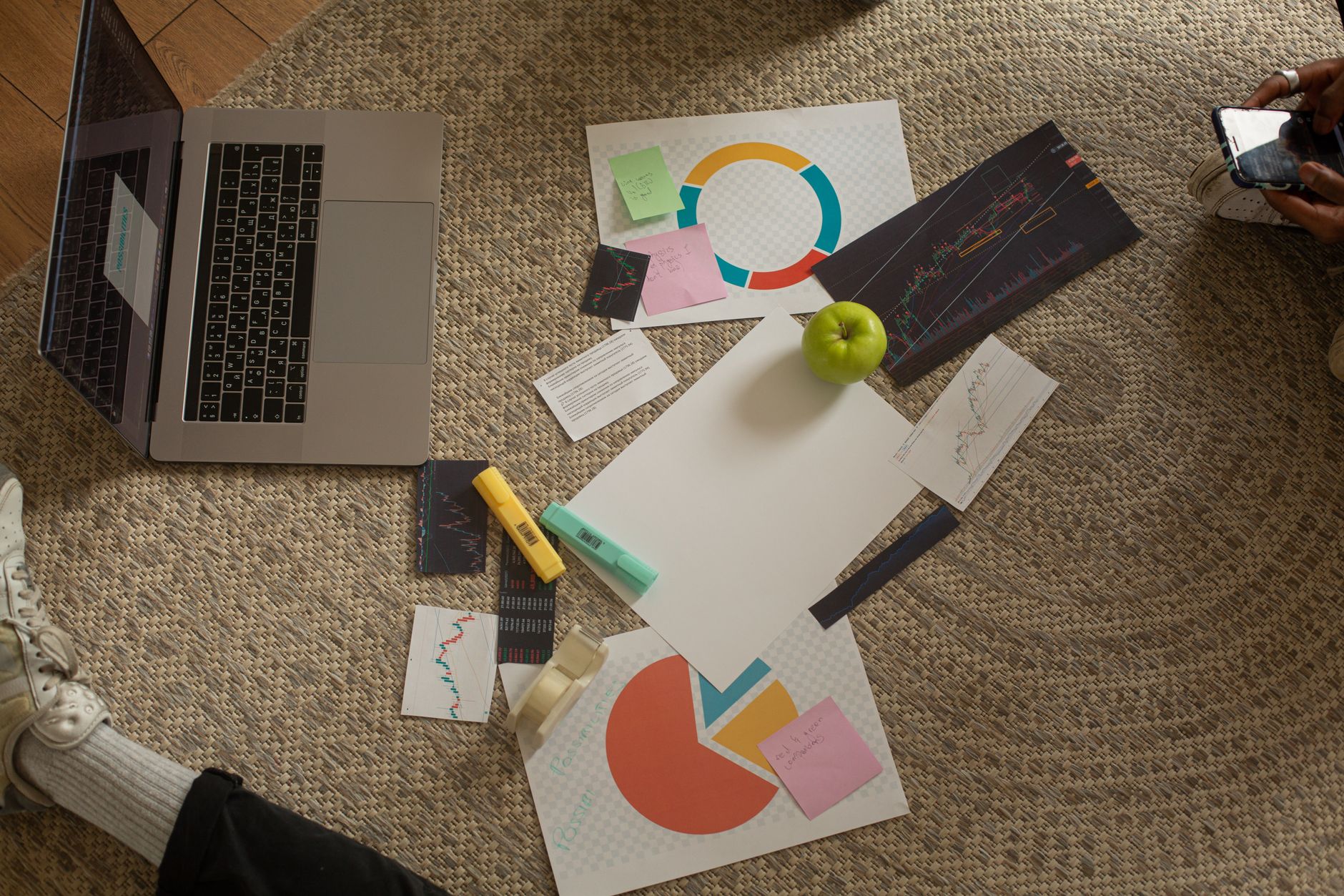 Top view of a workspace with a laptop, charts, sticky notes, and an apple on a woven rug floor.