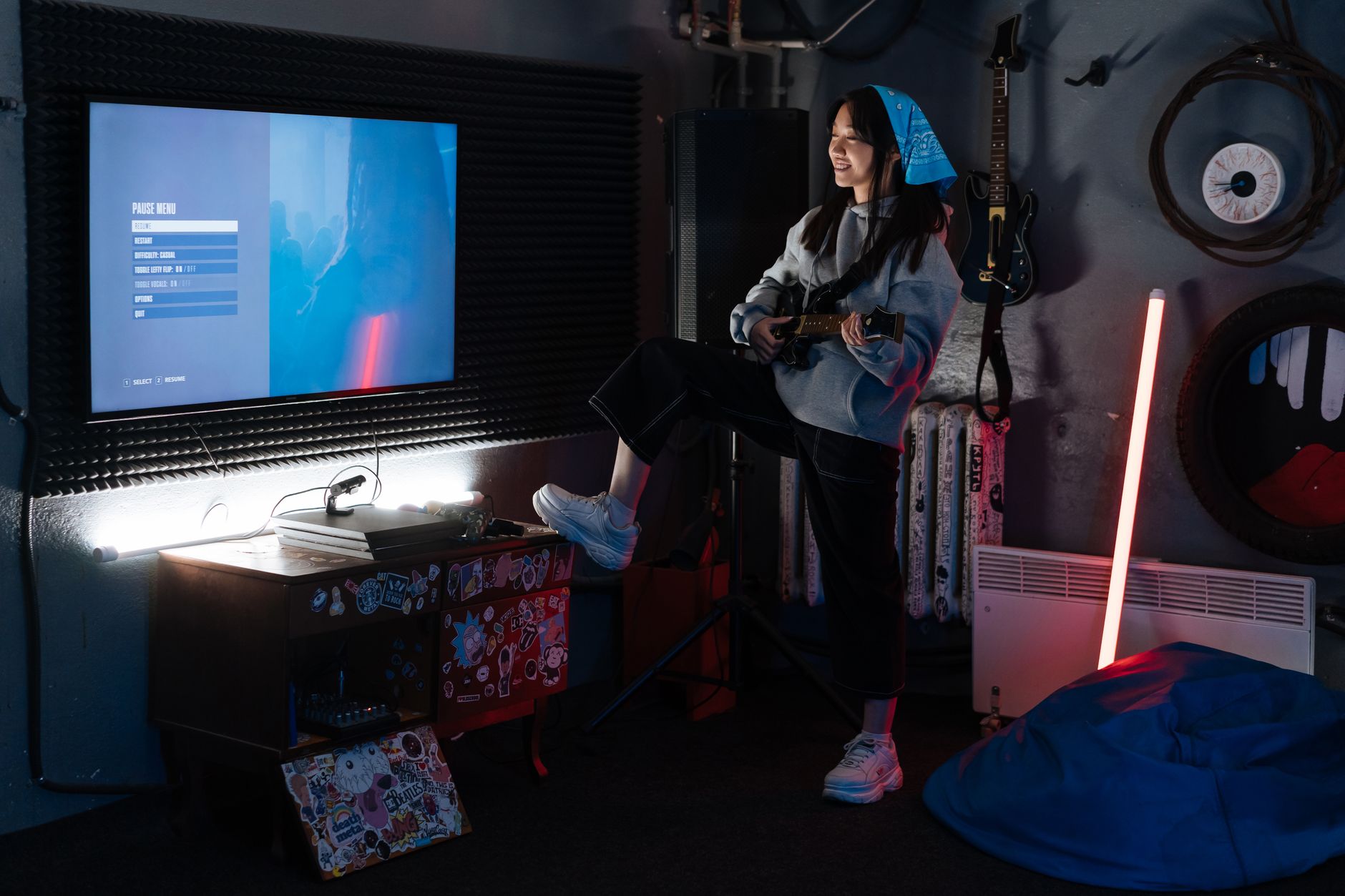 A young woman plays guitar in a stylish room with neon lighting and a wall-mounted screen.