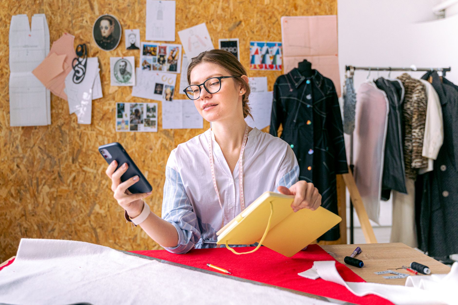 Fashion designer in studio using smartphone and notebook for creative work.