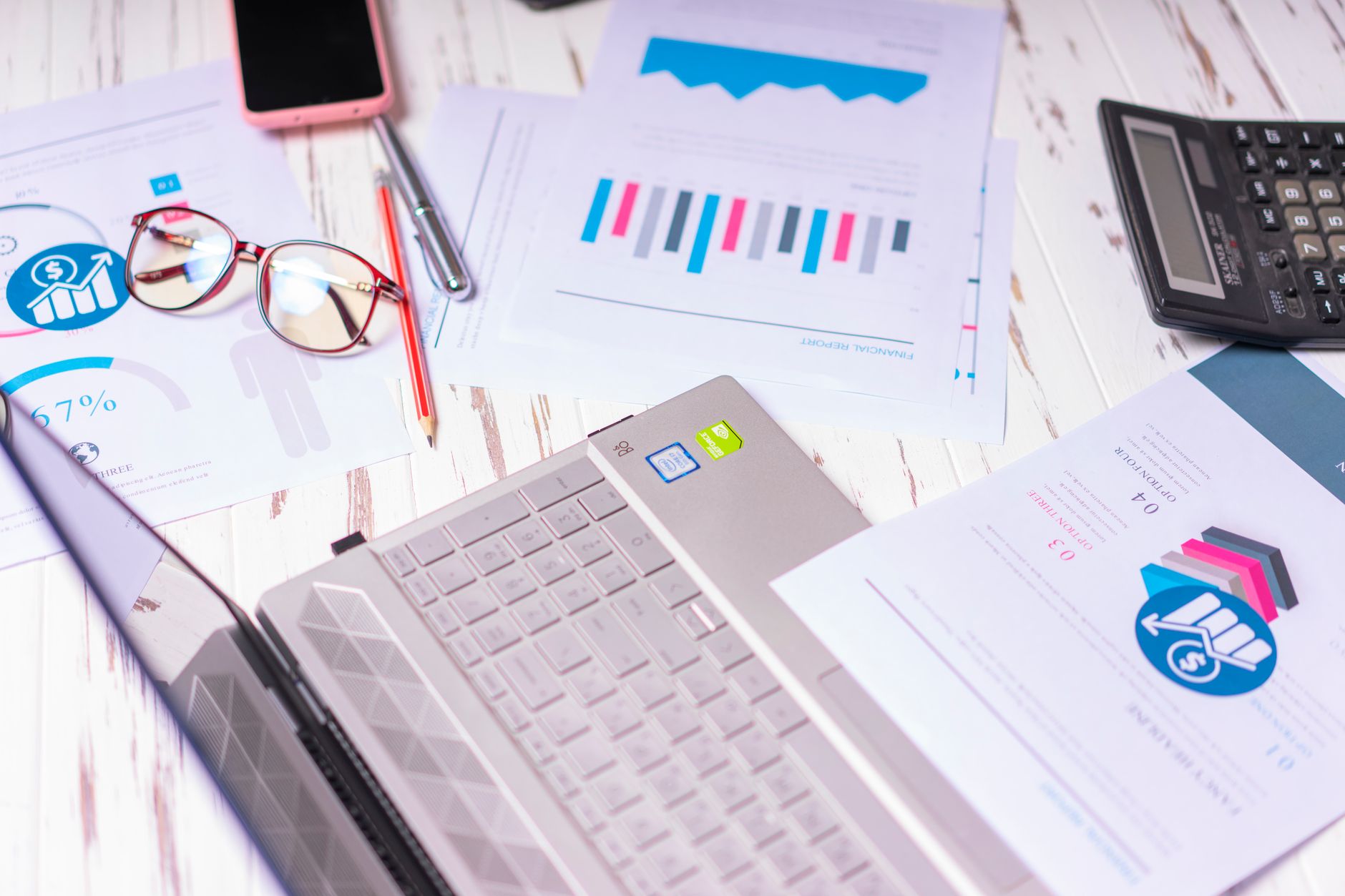 A cluttered desk with a laptop, financial reports, calculator, and glasses indicating a busy work environment.