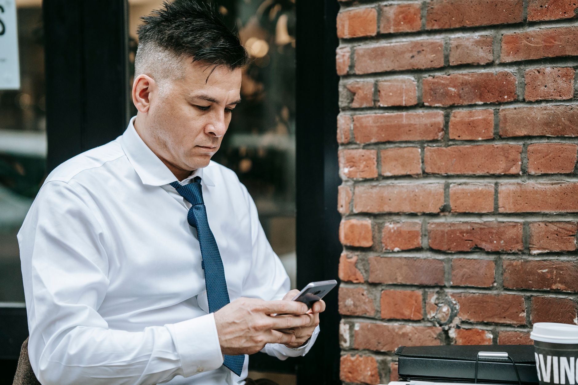 Professional man in corporate attire using smartphone outdoors by brick wall.