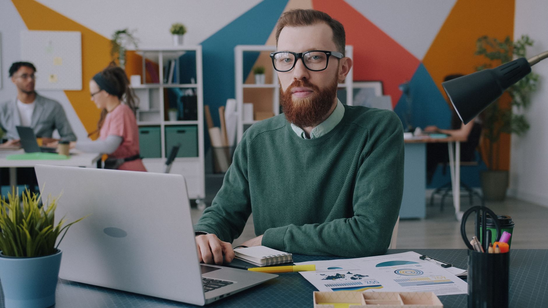 Man with glasses and beard working on laptop in colorful office.