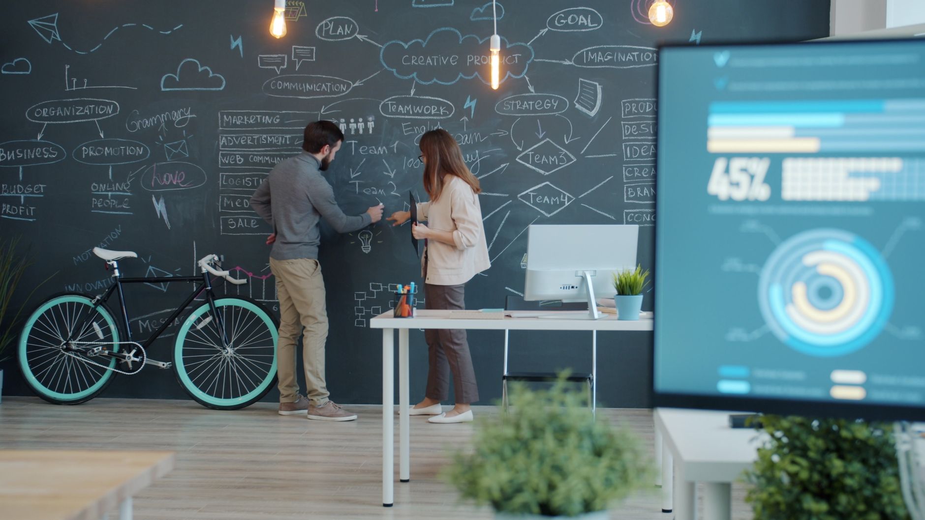 Two colleagues brainstorm ideas on a chalkboard in a modern office setting, discussing strategies and plans.