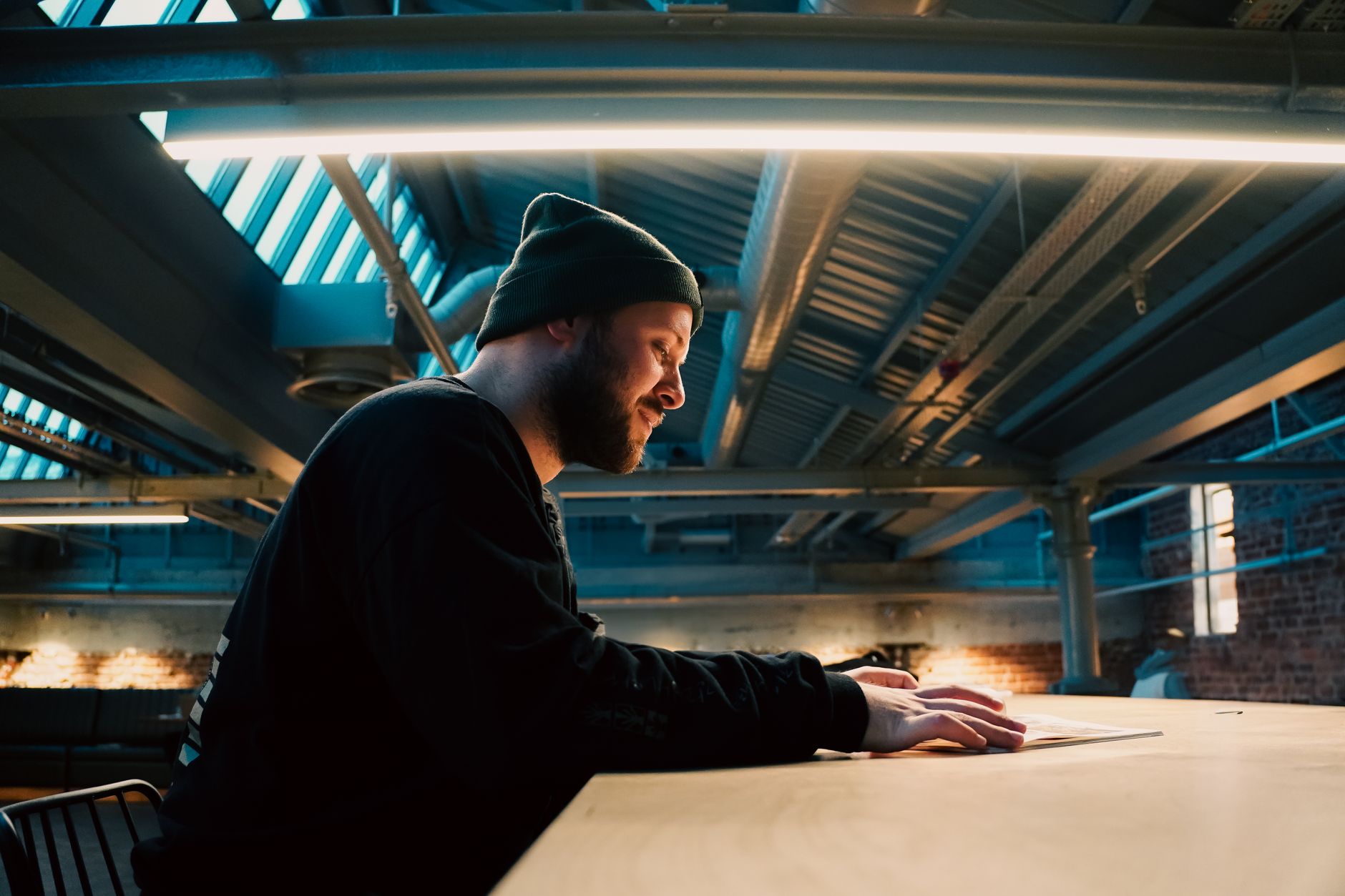 A man in a beanie reads under a modern industrial ceiling light in a loft setting.